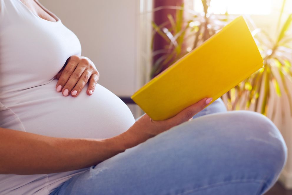 Eine schwangere Person hält eine Broschüre in der Hand, während sie eine Hand auf ihren Bauch gelegt hat