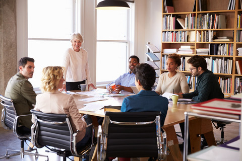 Eine mittelalte Frau steht an einem Tisch vor einer Gruppe sitzender, j&uuml;ngerer Personen